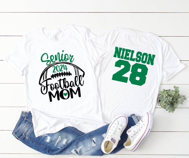 Op de afbeelding: Wit T-shirt met de tekst "Senior 2024 Football Mom" en een groen hart. Een tweede wit T-shirt met de tekst "NIELSON 28" in groen.