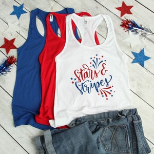 May include: Three tank tops in red, white, and blue. The white tank top has the text "Stars & Stripes" in red and blue glitter lettering with a red and blue firework design. The tank tops are folded and displayed on a white wooden surface.
