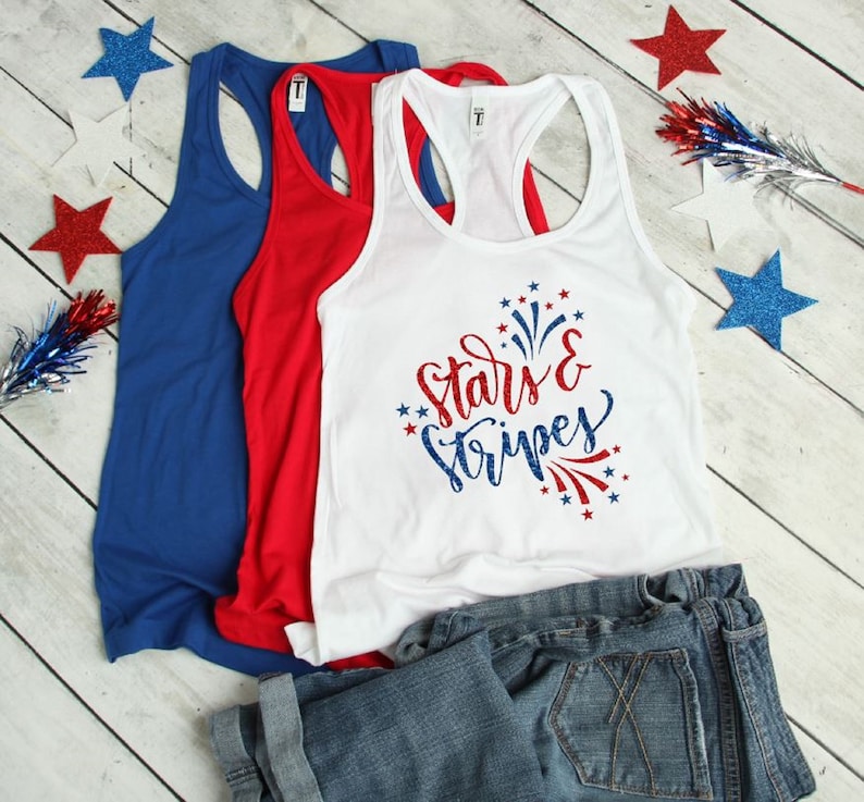May include: Three tank tops in red, white, and blue. The white tank top has the text "Stars & Stripes" in red and blue glitter lettering with a red and blue firework design. The tank tops are folded and displayed on a white wooden surface.