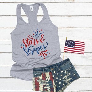 May include: A gray tank top with the text "stars & stripes" in red and blue glitter. The text is surrounded by red and blue stars and fireworks. The tank top is paired with denim shorts with a red, white, and blue American flag design.