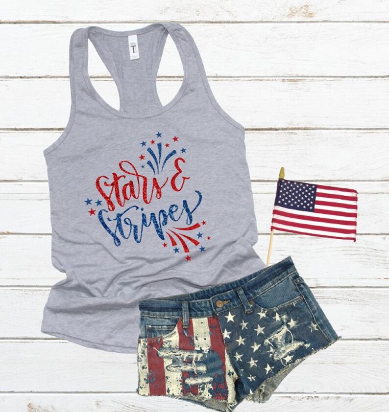 May include: A gray tank top with the text "stars & stripes" in red and blue glitter. The text is surrounded by red and blue stars and fireworks. The tank top is paired with denim shorts with a red, white, and blue American flag design.
