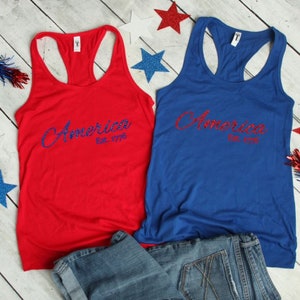 K&ouml;nnte beinhalten: Zwei Tanktops mit dem Wort "America" in blauen Glitzerbuchstaben und "Est. 1776" darunter. Die Tanktops sind rot und blau.