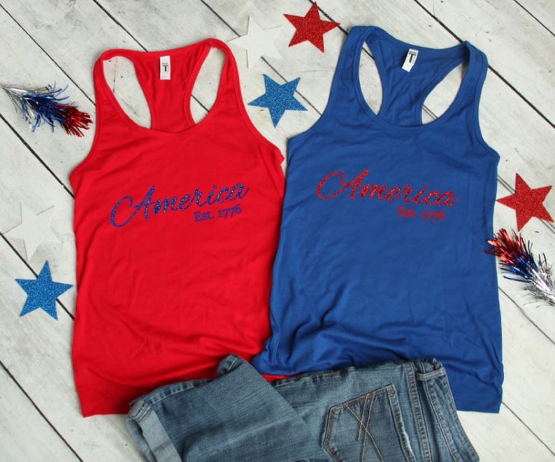 K&ouml;nnte beinhalten: Zwei Tanktops mit dem Wort "America" in blauen Glitzerbuchstaben und "Est. 1776" darunter. Die Tanktops sind rot und blau.