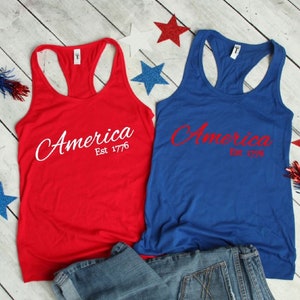 K&ouml;nnte beinhalten: Zwei Tanktops mit dem Text "America Est 1776" in Wei&szlig; auf einem roten Tanktop und in Rot auf einem blauen Tanktop. Die Tanktops liegen auf einer Holzoberfl&auml;che mit einer blauen Jeans.