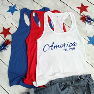 K&ouml;nnte beinhalten: Drei Tanktops in Blau, Rot und Wei&szlig;. Das wei&szlig;e Tanktop hat den Text "America Est. 1776" in Blau.