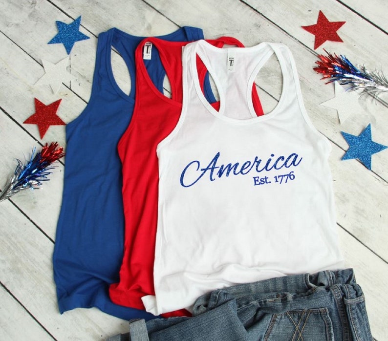 K&ouml;nnte beinhalten: Drei Tanktops in Blau, Rot und Wei&szlig;. Das wei&szlig;e Tanktop hat den Text "America Est. 1776" in Blau.