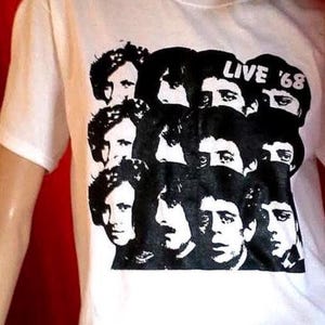 Pode incluir: Uma t-shirt branca de manga curta com uma impressão gráfica preta de vários rostos dispostos numa grelha. O texto "LIVE '68" está impresso a branco dentro da área gráfica preta. A t-shirt tem um decote redondo e um corte padrão.