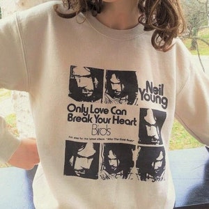 Op de afbeelding: Een crèmekleurige trui met een zwart-witte grafische print van de albumhoes van Neil Young voor "Only Love Can Break Your Heart". De albumhoes bevat een collage van zwart-witte afbeeldingen van Neil Youngs gezicht. De tekst "Only Love Can Break Your Heart" en "Birds" is in zwart op de trui gedrukt. De tekst "Neil Young" is in zwart op de rechterkant van de trui gedrukt.