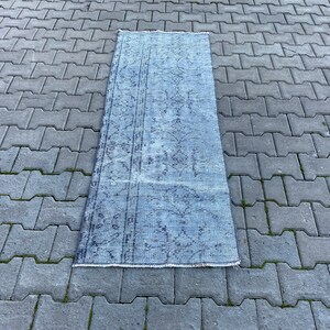 Puede incluir: Una alfombra rectangular azul claro con un aspecto descolorido y vintage. La alfombra presenta un patrón floral detallado en azul más oscuro y está colocada sobre una superficie de ladrillos grises. El diseño de la alfombra sugiere que es adecuada para un pasillo o entrada.