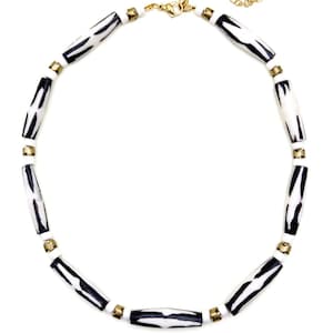 Peut inclure: Un collier composé de perles blanches et noires en forme de baril, avec des accents dorés. Le collier est doté d'une chaîne et d'un fermoir dorés. Les perles présentent un motif zébré. Le collier est présenté sur fond blanc.