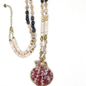 Peut inclure: Un collier à plusieurs rangs avec un grand pendentif en forme de coquillage rouge et blanc avec une bordure dorée. Le collier comprend diverses perles de couleur crème, noire et dorée, ainsi que de petits coquillages. La chaîne réglable est dorée.