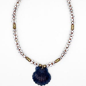 Peut inclure: Un collier de perles blanches et noires, entrecoupées d'accents dorés et de petites perles rouges. Le collier est orné d'un pendentif en forme de coquillage bleu foncé. Le fermoir est de couleur dorée.
