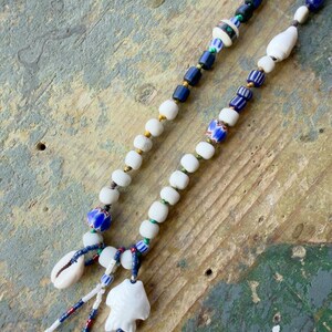 K&ouml;nnte beinhalten: Zwei Perlenketten mit wei&szlig;en, blauen und goldenen Perlen. Eine Halskette hat eine Muschel und einen wei&szlig;en Fischanh&auml;nger. Die andere Halskette hat eine Vielzahl von blauen und wei&szlig;en Perlen. Die Halsketten werden auf einer verwitterten, mehrfarbigen Holzoberfl&auml;che pr&auml;sentiert.