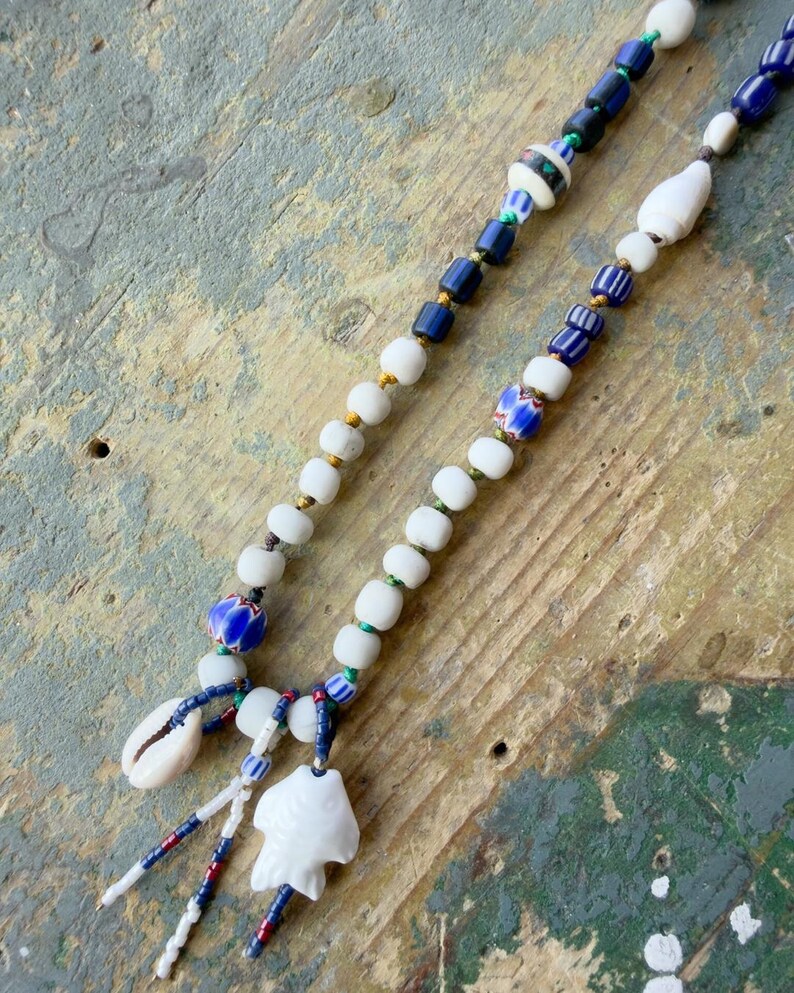 K&ouml;nnte beinhalten: Zwei Perlenketten mit wei&szlig;en, blauen und goldenen Perlen. Eine Halskette hat eine Muschel und einen wei&szlig;en Fischanh&auml;nger. Die andere Halskette hat eine Vielzahl von blauen und wei&szlig;en Perlen. Die Halsketten werden auf einer verwitterten, mehrfarbigen Holzoberfl&auml;che pr&auml;sentiert.