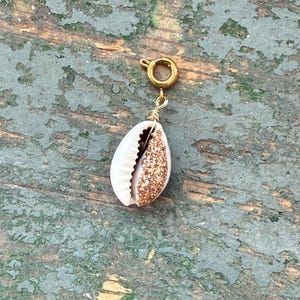 May include: A gold-colored charm with a white and glitter-filled seashell pendant. The seashell is attached to a gold-colored clasp. The charm is designed to be attached to a necklace or bracelet. The background is a weathered, green-painted surface.