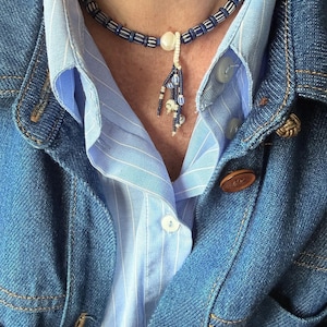 Peut inclure: Un collier de perles bleu et blanc. Le collier est composé d'une chaîne de perles bleues et blanches, avec un pendentif de perles blanches et d'accents bleus. Le collier est porté sur une chemise rayée bleue et une veste en jean.