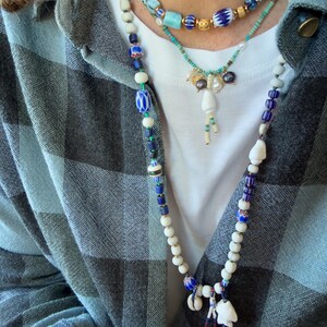 K&ouml;nnte beinhalten: Drei mehrreihige Halsketten mit verschiedenen Perlen. Die obere Halskette hat t&uuml;rkisfarbene und goldene Perlen. Die mittlere Halskette hat Perlen und einen Anh&auml;nger. Die untere Halskette hat wei&szlig;e, blaue und Muschelperlen. Die Person tr&auml;gt ein kariertes Hemd.