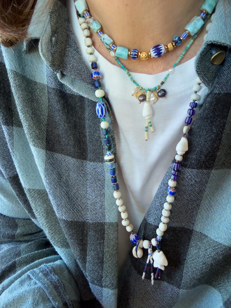 K&ouml;nnte beinhalten: Drei mehrreihige Halsketten mit verschiedenen Perlen. Die obere Halskette hat t&uuml;rkisfarbene und goldene Perlen. Die mittlere Halskette hat Perlen und einen Anh&auml;nger. Die untere Halskette hat wei&szlig;e, blaue und Muschelperlen. Die Person tr&auml;gt ein kariertes Hemd.