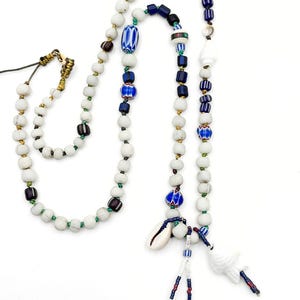 K&ouml;nnte beinhalten: Eine handgefertigte Halskette mit wei&szlig;en, blauen und braunen Perlen. Die Halskette hat einen Muschelanh&auml;nger und einen wei&szlig;en Keramikanh&auml;nger. Die Perlen sind mit gr&uuml;nen und goldenen Akzenten aufgef&auml;delt, wodurch ein einzigartiges und k&uuml;nstlerisches Design entsteht.