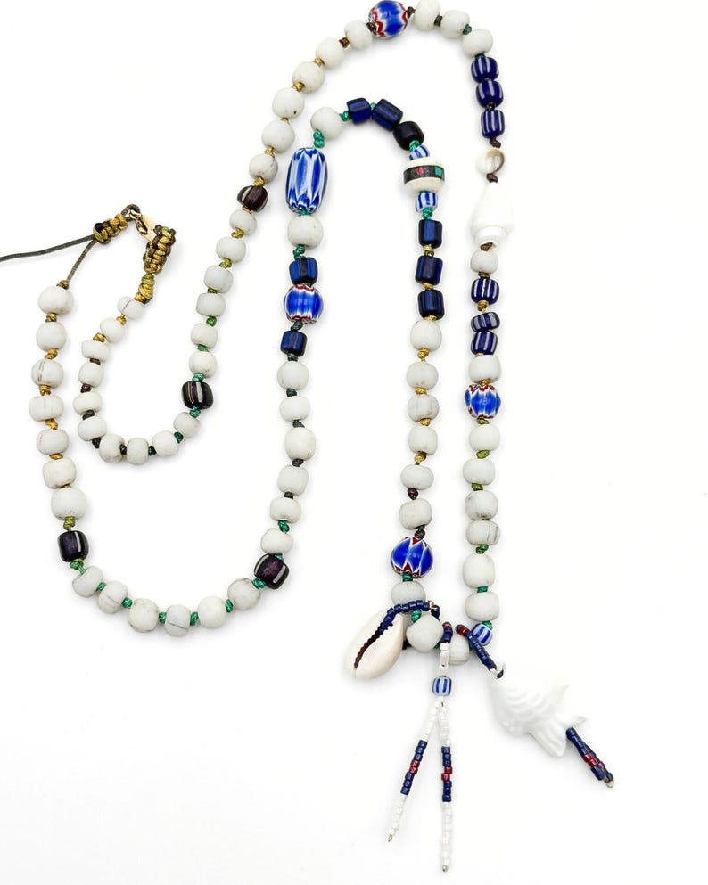 K&ouml;nnte beinhalten: Eine handgefertigte Halskette mit wei&szlig;en, blauen und braunen Perlen. Die Halskette hat einen Muschelanh&auml;nger und einen wei&szlig;en Keramikanh&auml;nger. Die Perlen sind mit gr&uuml;nen und goldenen Akzenten aufgef&auml;delt, wodurch ein einzigartiges und k&uuml;nstlerisches Design entsteht.