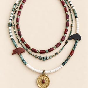 May include: A multi-strand beaded necklace featuring cylindrical red beads, small turquoise-colored stone chips, white disc beads, and metallic accents. The piece includes a carved brown bear charm, a black bear silhouette, and a round gold-toned pendant.