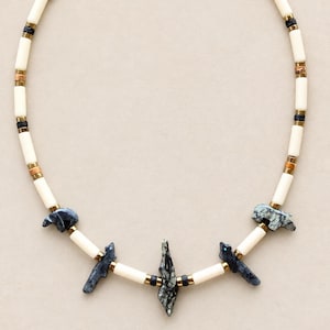 May include: A necklace featuring long white cylindrical beads, small metallic gold spacers, and dark textured stone beads. The design includes carved stone animal figures and a central pointed stone pendant, all arranged in a repeating pattern.