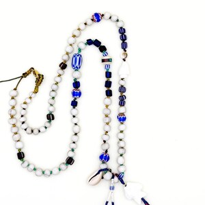 K&ouml;nnte beinhalten: Eine handgefertigte Halskette mit wei&szlig;en, blauen und schwarzen Perlen. Die Halskette enth&auml;lt einen Muschelanh&auml;nger und einen wei&szlig;en, abstrakt geformten Anh&auml;nger. Die Perlen sind mit dekorativen Elementen durchsetzt, was ein einzigartiges und k&uuml;nstlerisches Design ergibt.