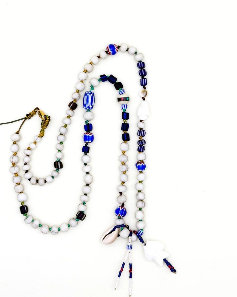K&ouml;nnte beinhalten: Eine handgefertigte Halskette mit wei&szlig;en, blauen und schwarzen Perlen. Die Halskette enth&auml;lt einen Muschelanh&auml;nger und einen wei&szlig;en, abstrakt geformten Anh&auml;nger. Die Perlen sind mit dekorativen Elementen durchsetzt, was ein einzigartiges und k&uuml;nstlerisches Design ergibt.