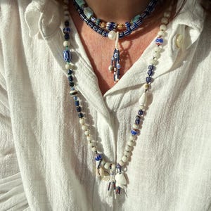 K&ouml;nnte beinhalten: Ein mehrschichtiges Halsketten-Set mit drei Str&auml;ngen aus Perlenketten. Die Halsketten umfassen wei&szlig;e, blaue und goldfarbene Perlen mit einem Anh&auml;nger aus kleinen Muscheln und Charms. Die Person tr&auml;gt ein wei&szlig;es Hemd mit Kn&ouml;pfen.