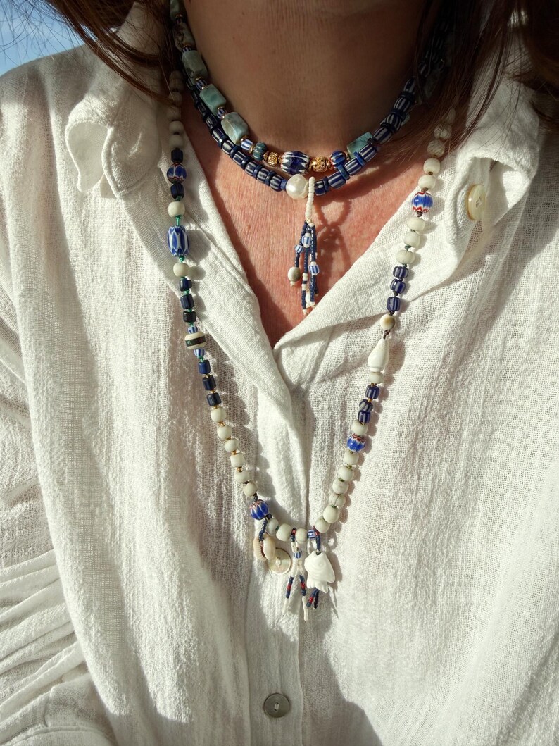 K&ouml;nnte beinhalten: Ein mehrschichtiges Halsketten-Set mit drei Str&auml;ngen aus Perlenketten. Die Halsketten umfassen wei&szlig;e, blaue und goldfarbene Perlen mit einem Anh&auml;nger aus kleinen Muscheln und Charms. Die Person tr&auml;gt ein wei&szlig;es Hemd mit Kn&ouml;pfen.