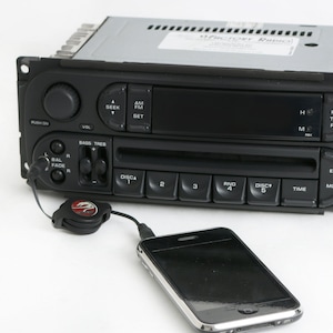 May include: A black car stereo with a CD player and a digital display. The stereo has buttons for AM/FM radio, seek, set, bass, treble, balance, fade, disc 1-5, random, eject, mode, and time. A black iPhone is connected to the stereo with a black aux cable.