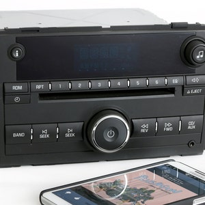 May include: A black car stereo with a digital display, buttons for radio stations, and a CD player. The stereo has a power button, seek buttons, and buttons for rewind and fast forward. A smartphone is in the foreground, displaying a music app.