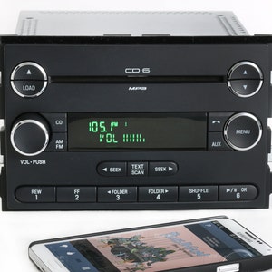 May include: A black car stereo with a digital display showing "105.1 FM VOL 111111". The stereo has buttons for CD, AM, FM, SEEK, TEXT SCAN, FOLDER, SHUFFLE, and OK. There are also buttons for LOAD, REW, FF, and MENU. The stereo is connected to a black smartphone.