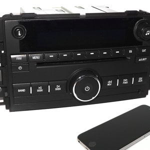 May include: A black car stereo with a digital display, buttons for radio functions, and a CD player. The stereo has a power button, seek buttons, and a band button. The stereo is connected to a black smartphone.