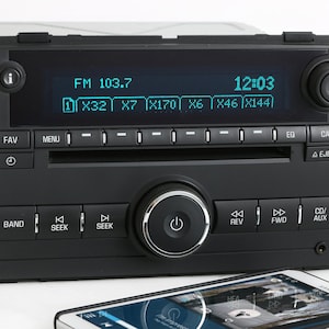 May include: A black car stereo with a digital display showing "FM 103.7" and "12:03". The stereo has buttons for "Fav", "Menu", "Band", "Seek", "EQ", "Cat", and "Eject".