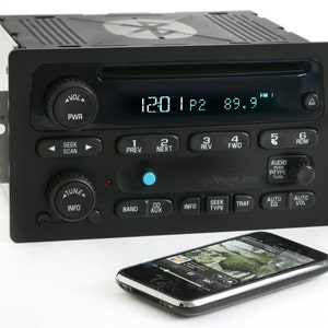 May include: Black car stereo with a digital display showing the time and radio frequency. Features buttons for power, seek, scan, and audio controls. A smartphone is placed in front of the stereo.