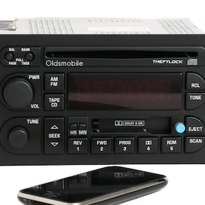 May include: A black car stereo with a silver faceplate and a display screen. The stereo has a variety of buttons and knobs, including power, volume, tune, seek, and eject. The stereo also has a cassette player and a CD player. The text "Oldsmobile" and "THEFTLOCK" is visible on the faceplate.