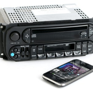 May include: A black car stereo with a CD player, cassette player, and radio tuner. The stereo has a digital display and several buttons for controlling the functions. A black smartphone with a white screen is connected to the stereo.