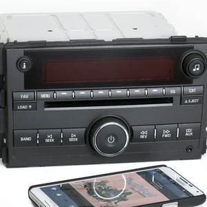 May include: A black car stereo with a CD player, radio tuner, and auxiliary input. The stereo has buttons for seek, band, load, eject, and other functions. The stereo is connected to a black smartphone with a screen displaying a music app.
