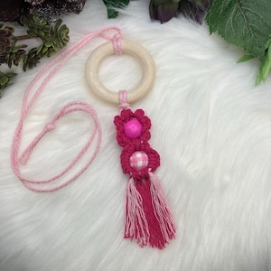 May include: A pink macrame car charm featuring a wooden ring and a decorative floral design. The charm includes a pink flower, a gingham bead, and a tassel. The charm is suspended by a pink cord.
