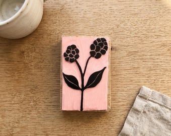 May include: A rectangular rubber stamp mounted on a wooden block featuring a black ink design of a plant with two stems, two pointed leaves, and two textured flower heads on a light pink background, positioned next to a ceramic mug.
