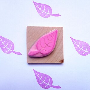 Op de afbeelding: Een roze rubberen stempel van een blad op een houten blok. De stempel is omringd door vier roze bladafdrukken op een witte achtergrond.