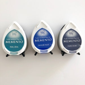 May include: Three ink pads in a teardrop shape with a white lid and a black base. The ink pads are labeled "Fade-resistant Dye Ink MEMENTO" with the colors Teal Zeal, Danube Blue, and Paris Dusk.