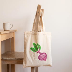 May include: A beige canvas tote bag featuring a hand-printed graphic of a purple beet with bright green leaves. The bag has two fabric handles and is displayed hanging from the back of a wooden chair next to a ceramic mug on a table.