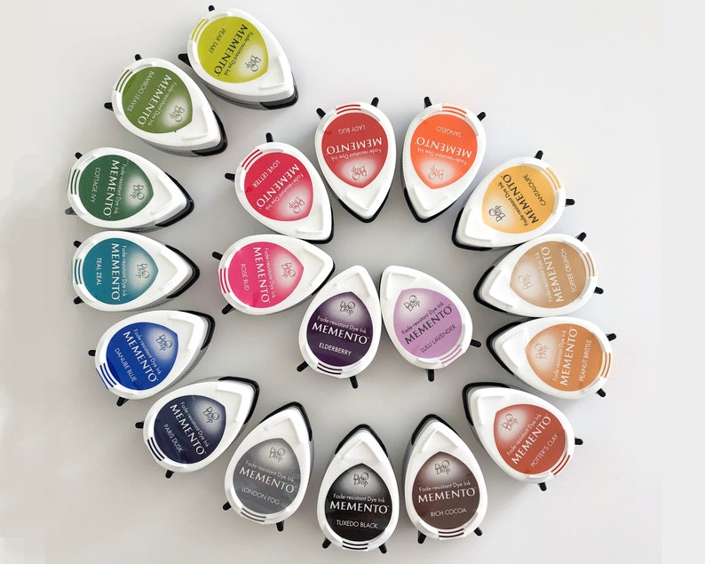 May include: A collection of 16 Memento Ink Pads in various colors, arranged in a circle. The ink pads are shaped like teardrops and have a white label with the brand name and ink color printed on them.