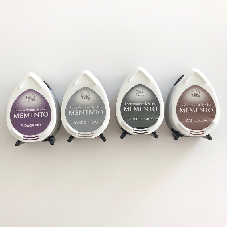 May include: Four ink pads for stamping, each with a different color ink. The ink pads are labeled "Dew Drop Fade-resistant Dye Ink Memento" with the color name below. The colors are Elderberry, London Fog, Tuxedo Black, and Rich Cocoa.