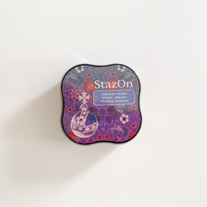 K&ouml;nnte beinhalten: Ein rundes, violettes Stempelkissen mit einem floralen Design und dem Text "StazOn Vibrant Violet Violet vibrant Violeta intenso" in Wei&szlig;.