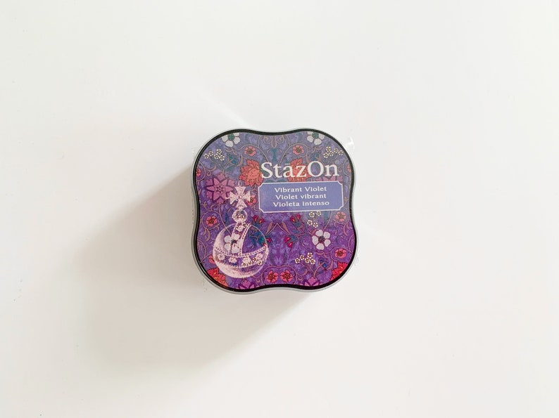 K&ouml;nnte beinhalten: Ein rundes, violettes Stempelkissen mit einem floralen Design und dem Text "StazOn Vibrant Violet Violet vibrant Violeta intenso" in Wei&szlig;.