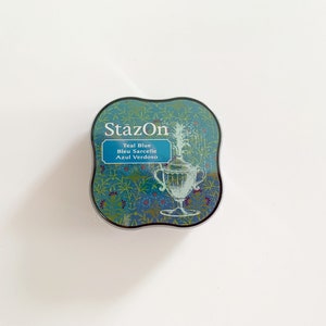 K&ouml;nnte beinhalten: Ein rundes, blaugr&uuml;nes Stempelkissen mit silbernem Rand. Das Stempelkissen hat ein Blumenmuster und den Text "StazOn Teal Blue Bleu Sarcelle Azul Verdoso" aufgedruckt.