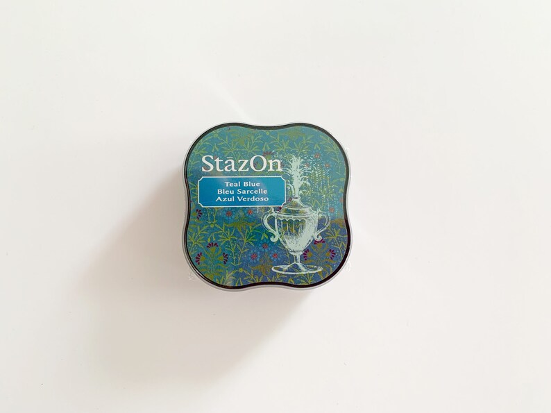 K&ouml;nnte beinhalten: Ein rundes, blaugr&uuml;nes Stempelkissen mit silbernem Rand. Das Stempelkissen hat ein Blumenmuster und den Text "StazOn Teal Blue Bleu Sarcelle Azul Verdoso" aufgedruckt.